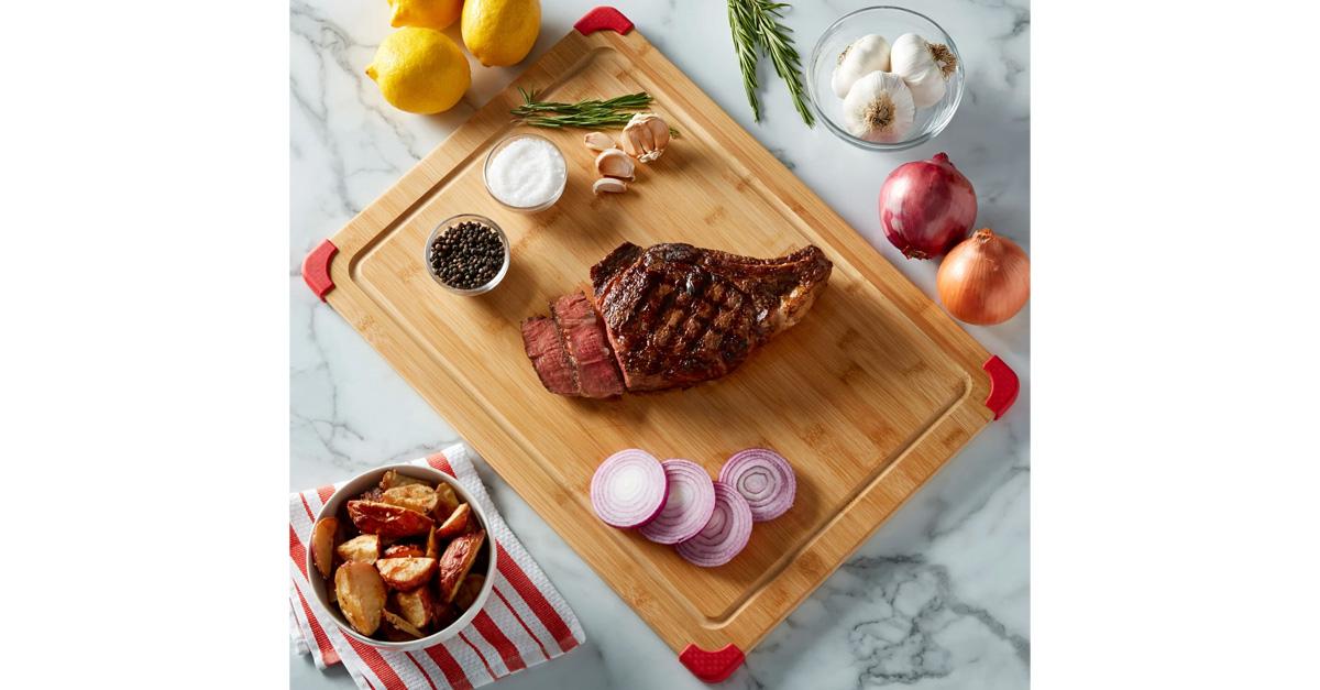 Bamboo cutting board with sliced steak, fruits, vegetables, and potatoes on a marble counter