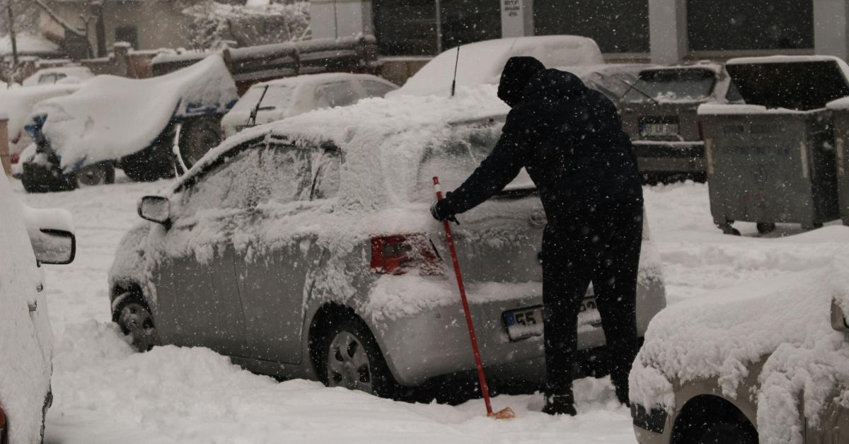 Person dressed in all black snow gear digging out a silver car trapped in snow. 