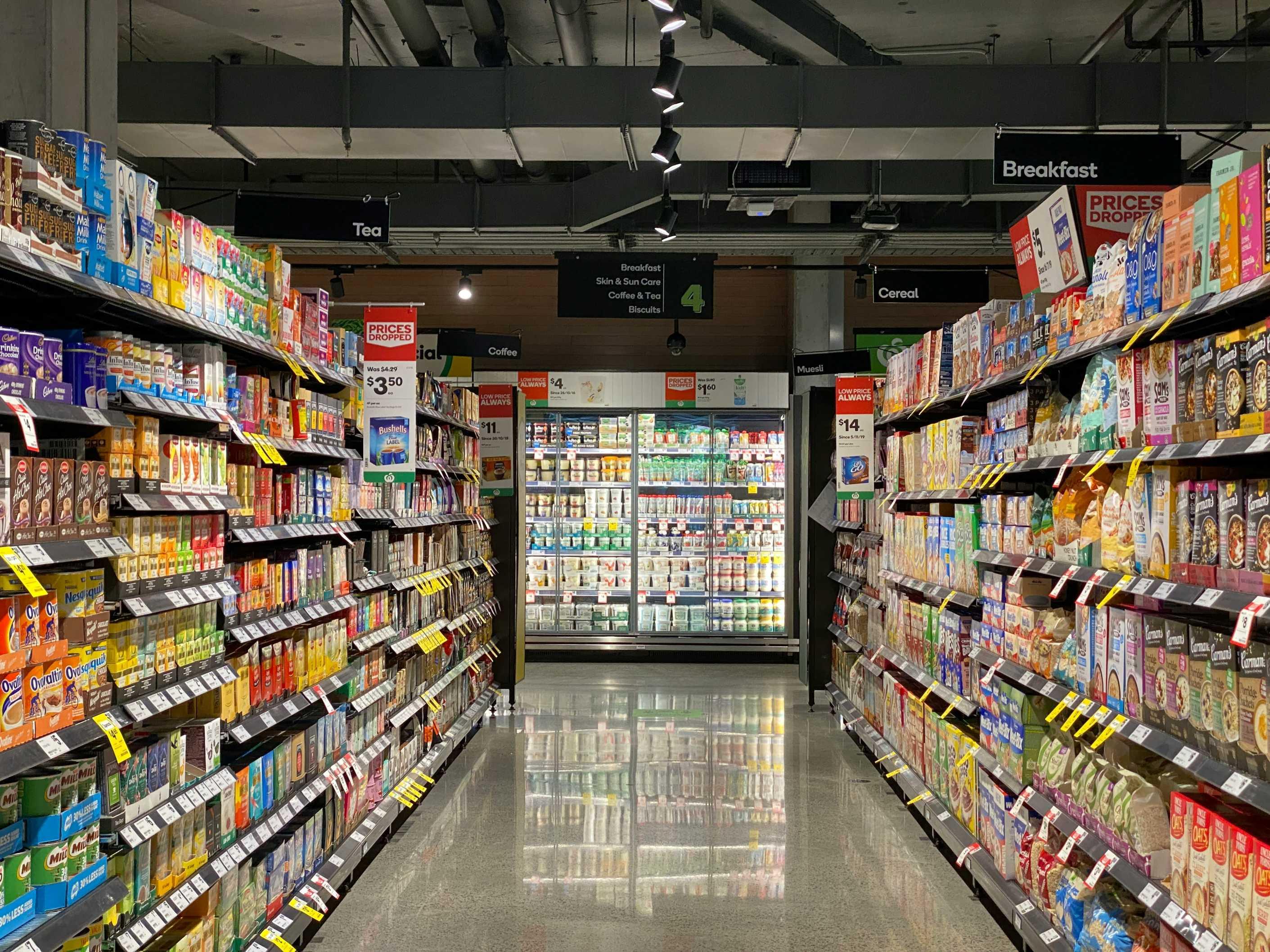 A grocery store aisle is pictured, featuring shelf-stable boxes and cans of food, in front of a refrigerated foods section.