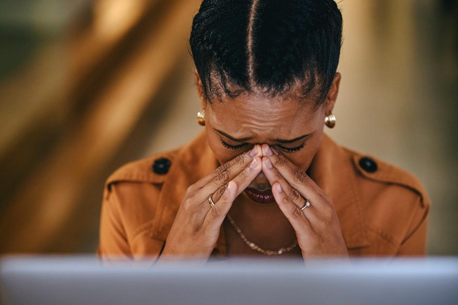 A woman wearing a brown jacket looks down with a pained expression while pressing her fingers to her nose. 