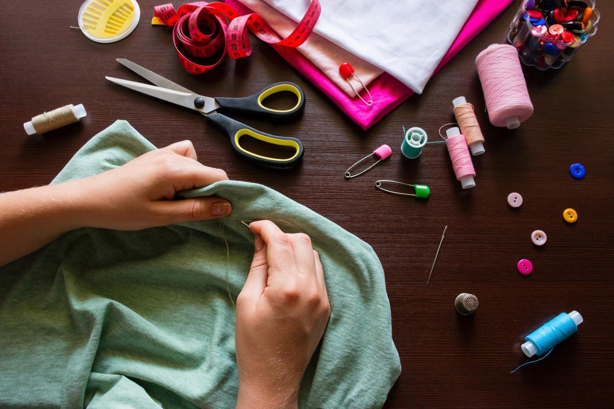 A person hand sewing a green piece of fabric on a dark wood table surrounded by colorful sewing supplies.