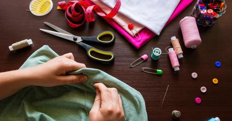 A person hand sewing a green piece of fabric on a dark wood table surrounded by colorful sewing supplies.