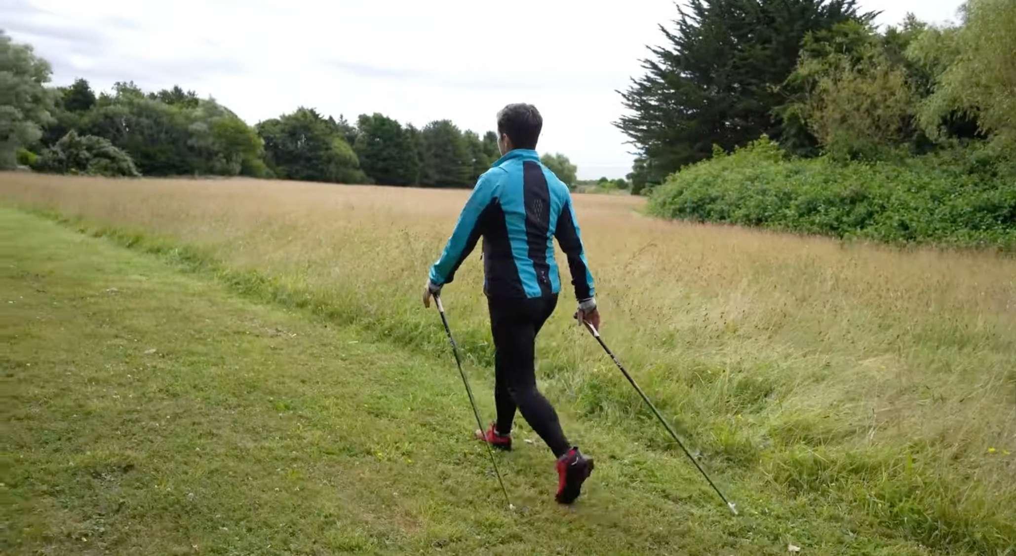 A woman walks across a grassy field using two walking poles.