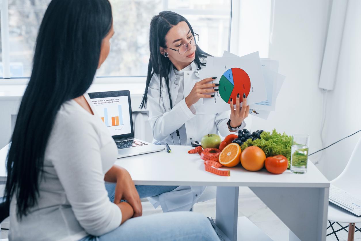 A dietitian explains healthy eating to a client while holding a chart beside a laptop and fruits and vegetables.