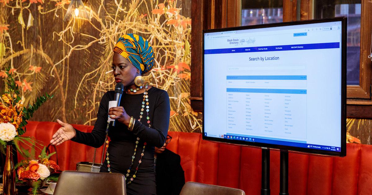 Nicole Deggins speaks into a microphone next to a screen showing the Black Doula Directory