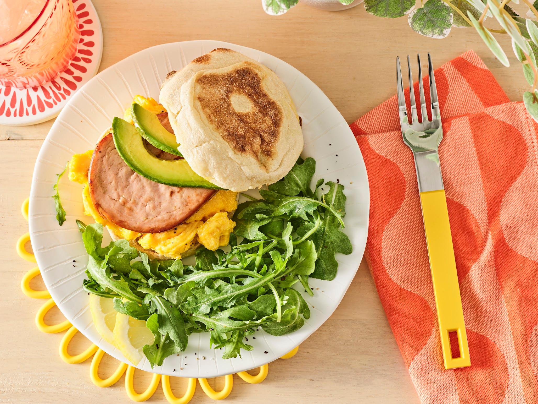 A Meati breakfast patty is pictured atop vegan eggs, along with avocado slices, on an English muffin, beside arugula.