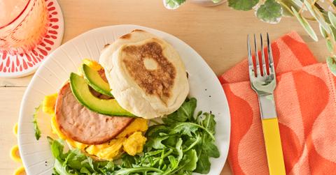 A Meati breakfast patty is pictured atop vegan eggs, along with avocado slices, on an English muffin, beside arugula.