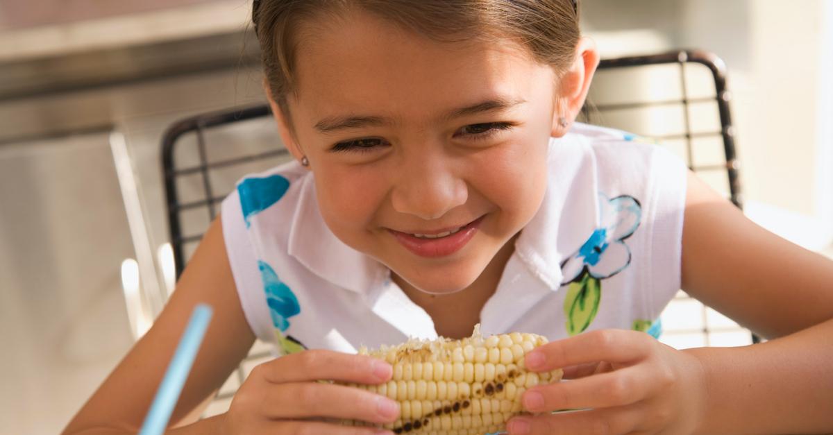 Child smiles and eats corn on the cob.