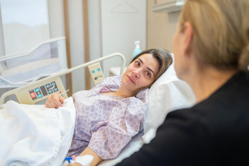 An over the shoulder shot of a woman in a hospital bed looking at a nurse with a sorrowful expression on her face. 